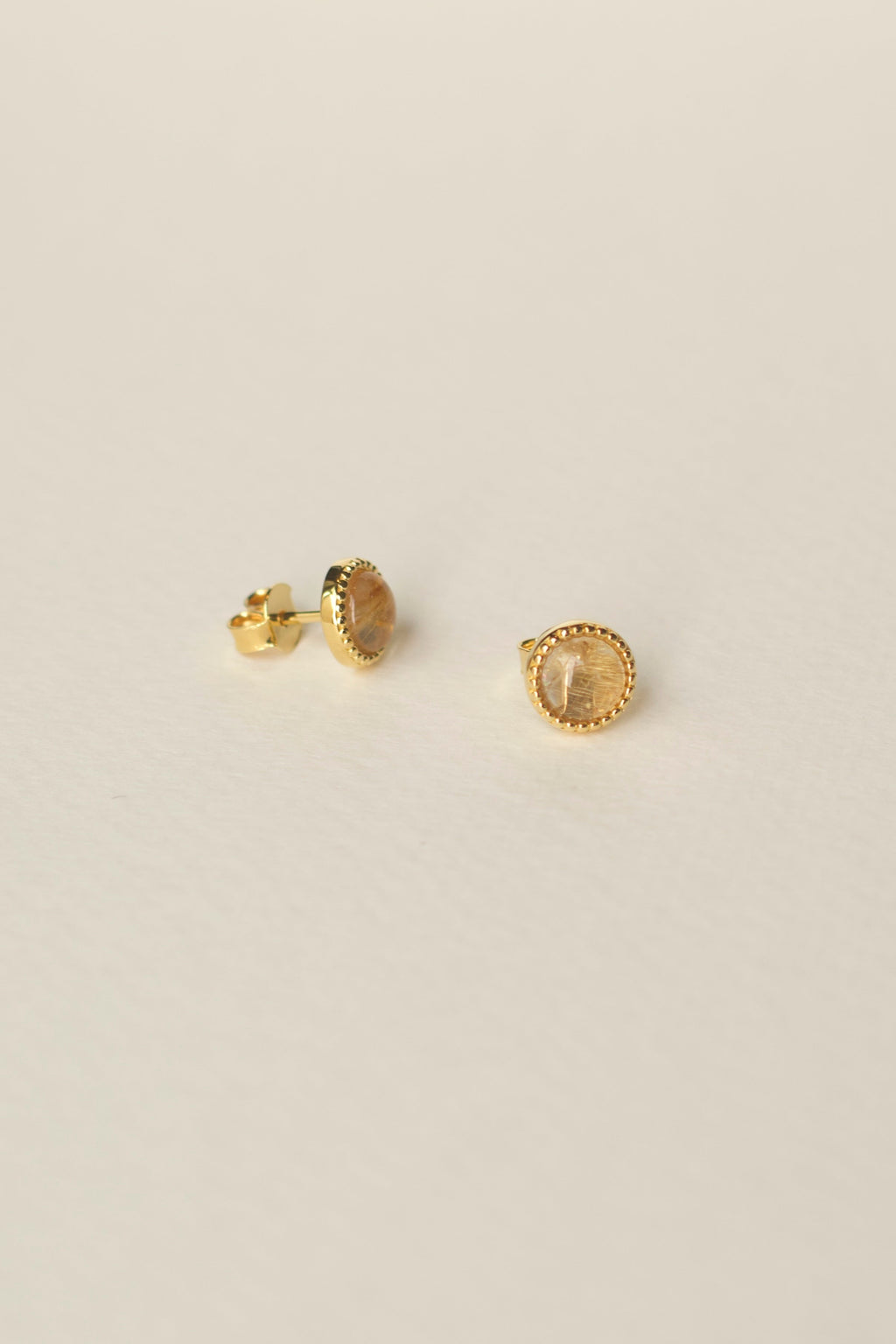 Pair of gold stud earrings with yellow rutilated quartz gemstone on a light beige background