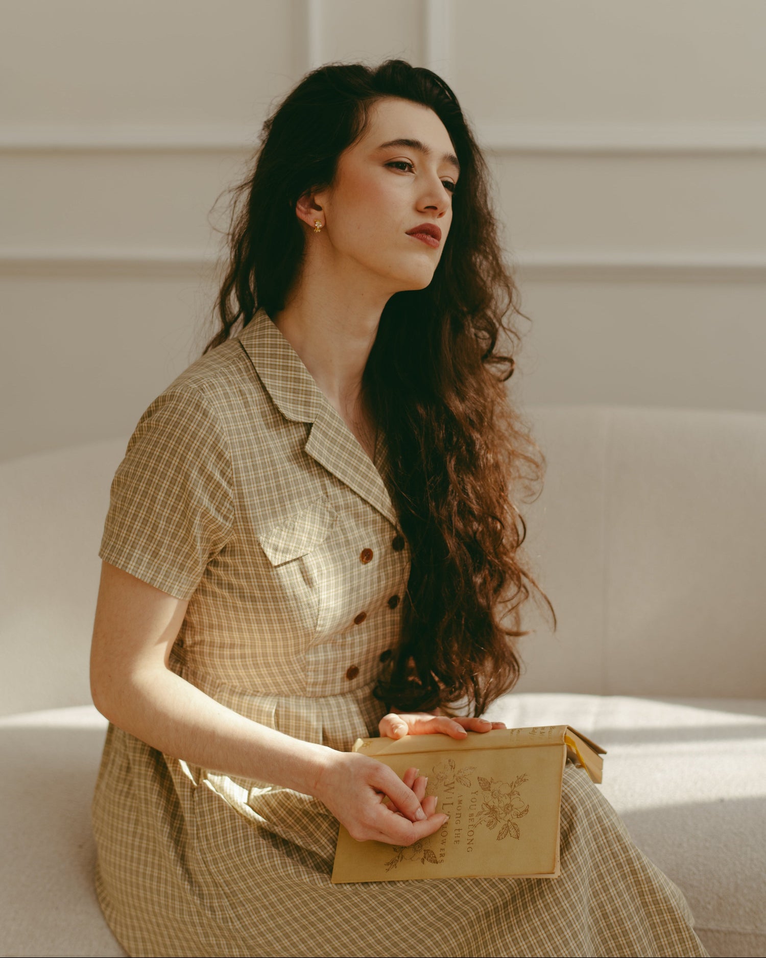 Woman in a checkered dress holding a book in a softly lit room.