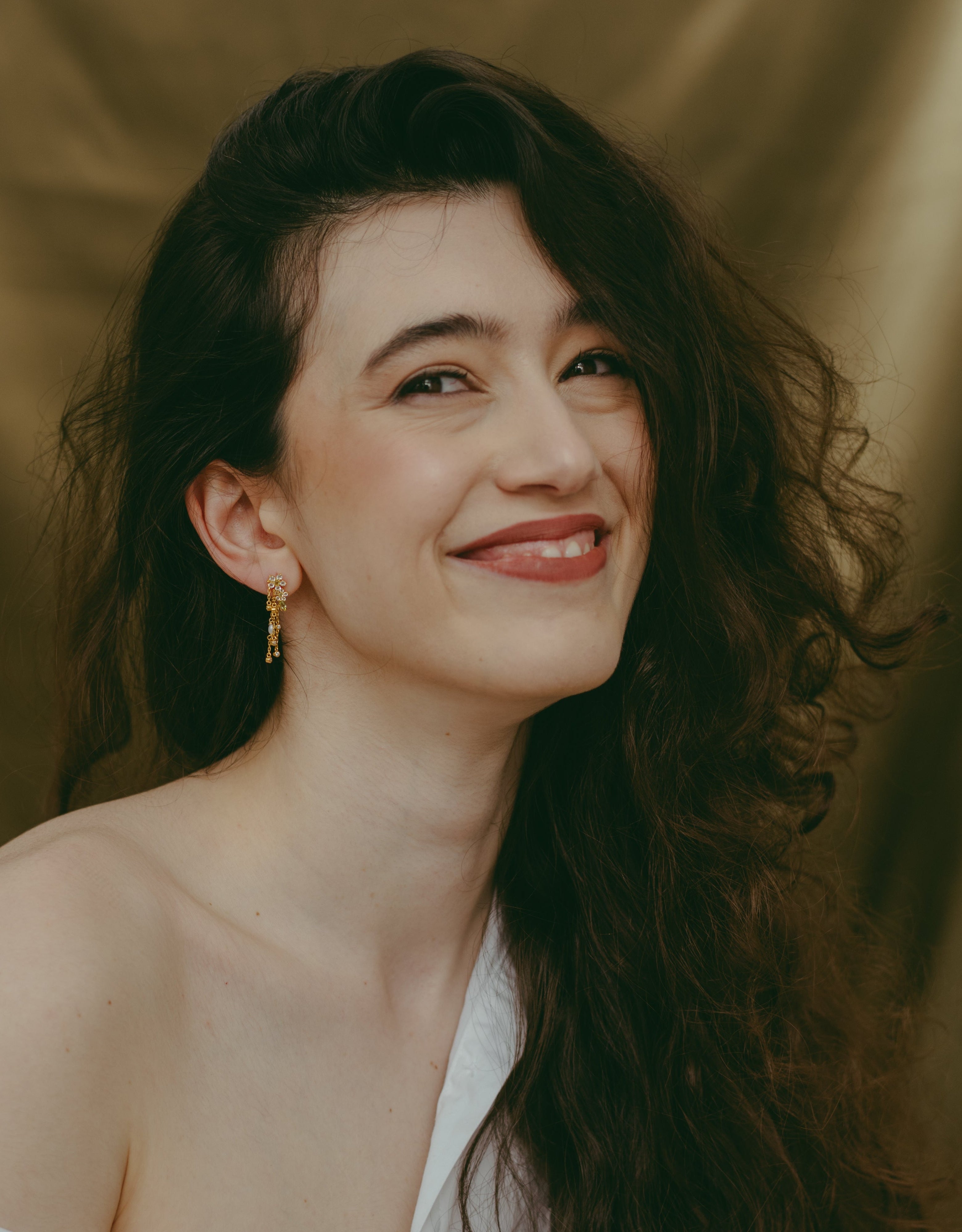 Woman with long dark hair wearing a white off-shoulder top against a gold curtain background